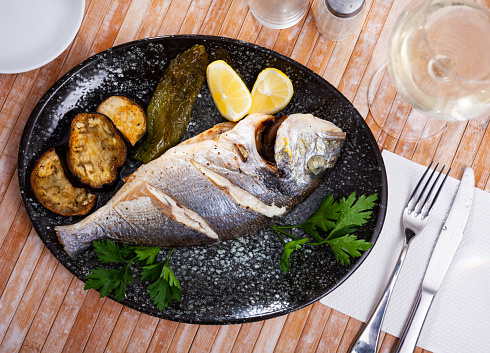 Appetizing grilled dorada fish served with vegetable garnish of baked eggplant and sweet pepper, lemon slices and greens