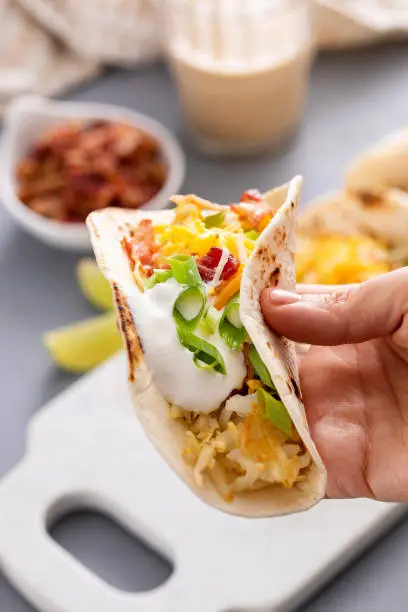Breakfast tacos with hashbrowns, scrambled eggs and bacon topped with cheese and green onion, hand holding a taco