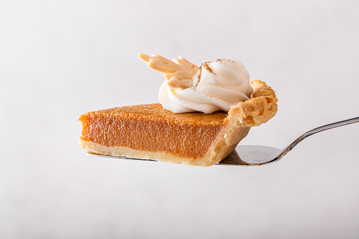 Slice of traditional pumpkin pie topped with whipped cream being held on a spatula on light background