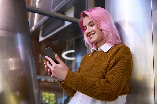 Portrait of young stylish woman standing in room, holding mobile phone and looking at the screen