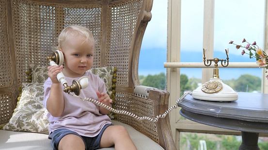 A cute little blond girl of two years, sits in a chair woven from a vine. She is holding the handset of an antique wire telephone, then brings it to her ear to hear the speaker.