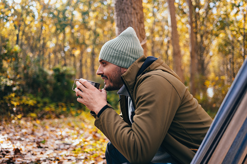 Smiling man sitting in a hammock and drinking cup of tea while camping in the nature.