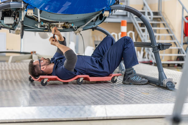 istock Aviation engineer lying on cart on wheels and fixing helicopter engine with work tool, wide shot 1444003821