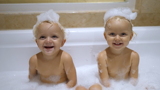 Two white identical girls take a bath with an abundance of foam. Foam party before bedtime in the bathroom. Creativity fun swimming for kids. The concept of hygiene and healthcare.