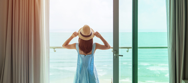 Happy woman wearing blue dress and hat looking outside window to ocean view in morning. Relax, vacation, time to travel and freedom concept