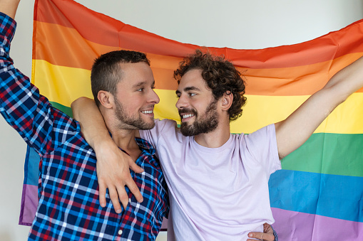 Two young gay lovers looking at each other affectionately. Two young male lovers standing together against a pride flag. Affectionate young gay couple sharing a romantic moment together.