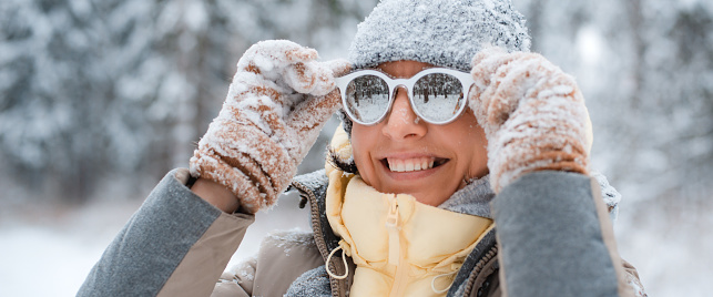 Happy woman in warm clothes, knitted hat and sunglasses standing outdoors during heavy snowfall, female watching falling snowflakes in park and rejoicing frosty winter weather. Selective focus