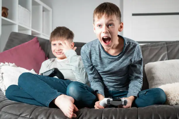 Portrait of two excited teenage boys children sitting on grey sofa at home with open mouth, holding gaming controller joystick, playing videogames, shouting. Hobby, free time, gaming, overemotive.