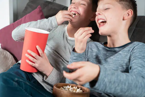 Photo of Portrait of two cheerful teenage boys sitting on sofa at home, watching funny movie on TV eating popcorn from red bucket