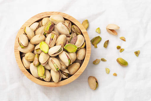 Pistachio nuts with shells in wooden bowl on white linen background, top view
