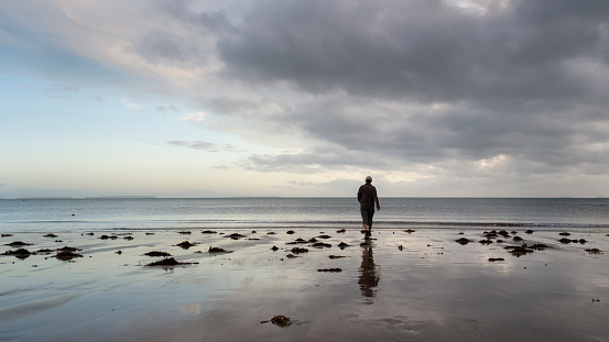 Silhouette man walking on the beach towards the sea, moody cloudy sky overhead.