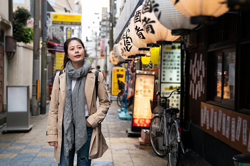 cheerful Asian Taiwanese woman traveler looking at the hanging lanterns outside a Japanese bar while walking leisurely on food street in Shinsekai area in Osaka Japan. Translation: alcoholic drinks