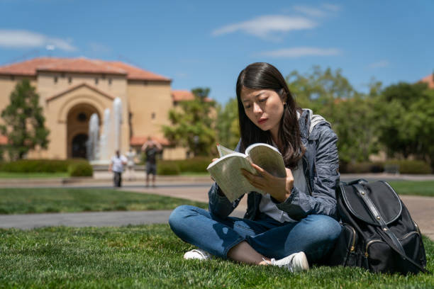 istock asian Japanese girl exchange student turning pages with serious face while reading a difficult book on grass in front of Memorial Auditorium Fountain at school university in California usa 1444104019