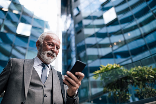Low angle view of smiling senior entrepreneur socializing on smart phone. Businessman wearing suit is carrying laptop bag. He is standing against office building in the city.
