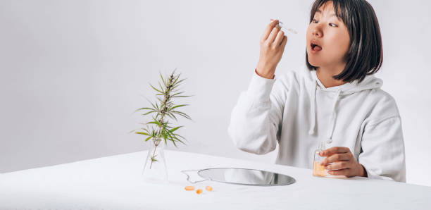 Asian woman dripping cbd oil into her mouth. Pharmaceutical medicine pills, capsules on a white table. Concept Melatonin production and restore sleeping routine. stock photo