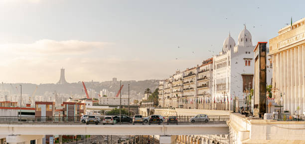 Algiers, Algeria - October 14, 2022: People's National Assembly and Wilaya headquarters building with birds flying, Arab league countries flags and the Martyr's Memorial monument in a coudy blue sky with diffused sunlight background.