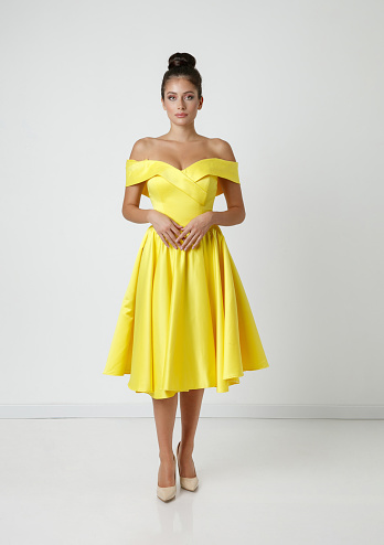 Beautiful young woman with brown hair in bun wearing yellow dress off shoulder and looking at camera, studio shot, beauty and fashion industry