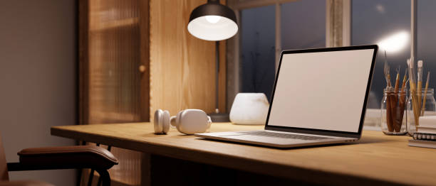 istock Cozy Scandinavian home workspace with laptop mockup, light from table lamp on wood table. 1444114847