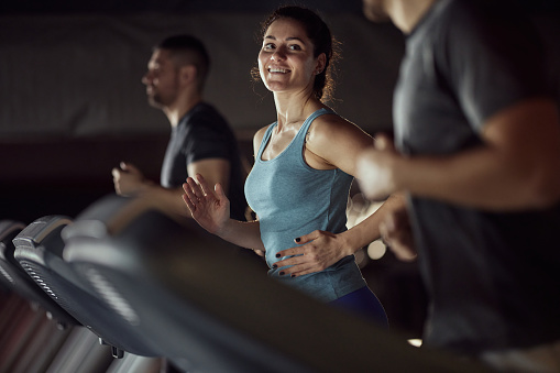 Happy female athlete taking to her friend while running on treadmill during sports training in a health club.