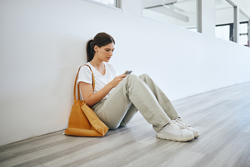 Sad university woman, student alone on floor in the hallway after class or lecture and browsing social media on a smartphone. Depression, stress and anxiety from college academic pressure for success