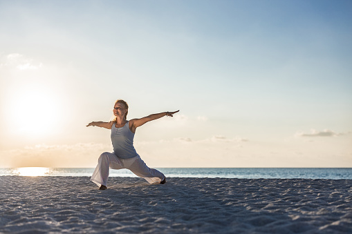 Smiling woman doing Yoga balance exercises with her arms outstretched on the beach at sunrise. Copy space.