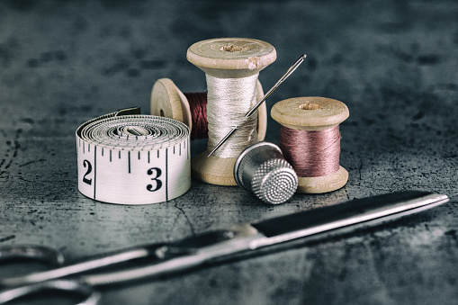 Spools of thread of different colors with a pin pinned together with a sewing scissors, a yellow tape measure, a brown button and a metal thimble on a white background.