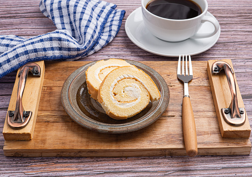 Top view of roll cake on a plate on a wooden tray with a cloth and a white coffee cup on a wooden table
