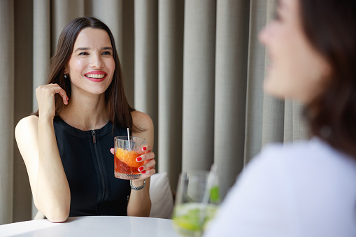 Beautiful young woman smiling and talking to her friend, sitting together at bar table and having drinks