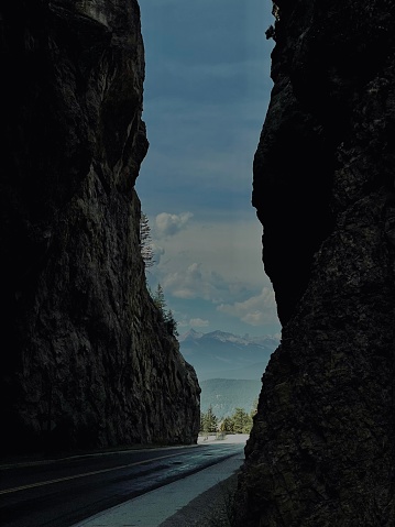 Two imposing shear vertical rock faces guard an empty road leading around a gentle bend. In the distance, deep woodlands and vast mountains give way to a poignant gorgeous blue sky and dramatic clouds.