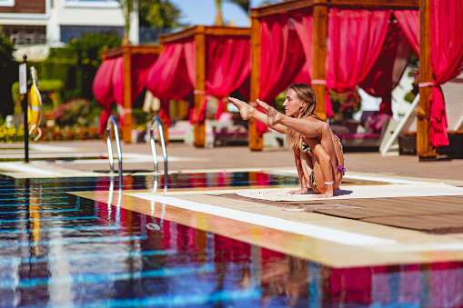 Woman doing yoga by the resort pool, balancing on arms with legs straight, feet extended up - Tittibhasana or Firefly pose
