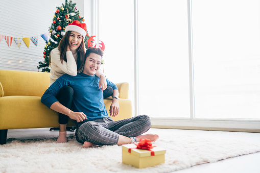 Young beautiful happy Asian woman wearing Santa Claus hat surprises and laughs in the back of her boyfriend at home with a Christmas tree in the background. Image with copy space.