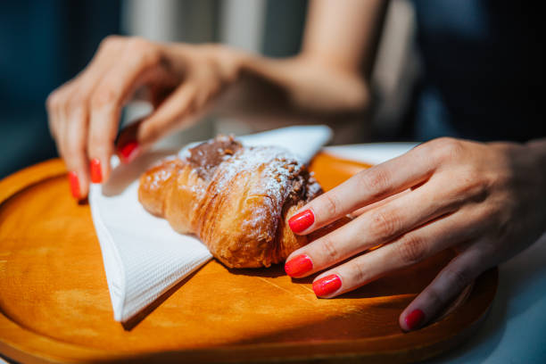 Female hands taking chocolate croissant with powdered sugar icing from wooden board, indulging in little luxuries