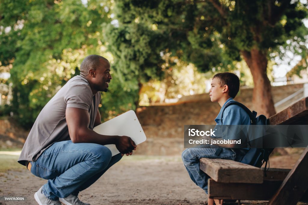 Mentor talking to a young school kid outside class Mentor talking to a young school kid outside class. Primary school teacher motivating a young boy. Teacher providing support and encouragement for a pupil in elementary school. Child Stock Photo