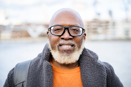 Head and shoulders view of bald man with gray facial hair wearing eyeglasses, orange sweater, and gray fleece jacket. Defocused cityscape in background.