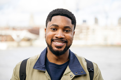Headshot of mid 20s man with short hair, beard, and mustache wearing khaki and navy blue warm clothing and smiling at camera. Defocused cityscape in background.