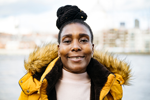 Woman with locs topknot wearing pastel mock turtleneck sweater, yellow parka with faux fur collar, and smiling at camera. Defocused cityscape in background.