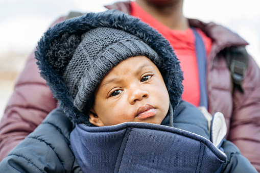 Close-up of young woman standing outdoors and supporting infant son in carrier as he looks at camera with curiosity.