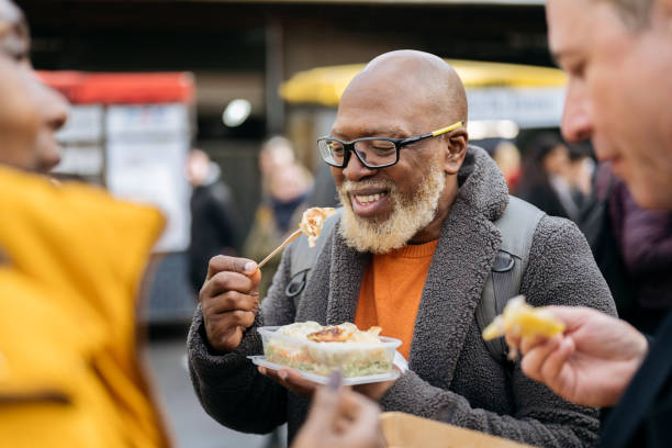 Smiling Londoner enjoying convenience food outdoors Mature Black couple and Caucasian friend talking and eating as they spend weekend leisure together at Southwark market hall. borough market stock pictures, royalty-free photos & images