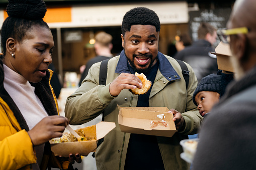 Waist-up view of smiling young Black man with baby boy and grandparents standing outdoors talking and eating specialty food in Southwark.