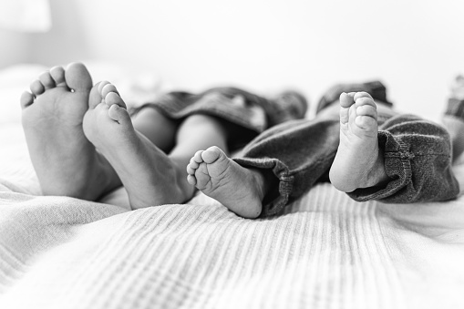 Black and white close-up of the feet of a newborn baby and it's sibling lying next to each other on a bed with white linen