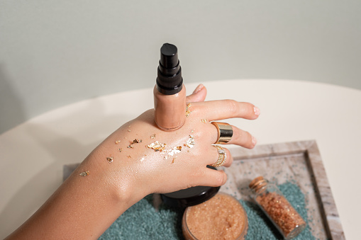 Unrecognizable woman hand with golden sparkle tinsel hold tinting cream, liquid make up foundation on back of the hand above tray with powder and granule bath salt. Cosmetics and beauty. Advertisement