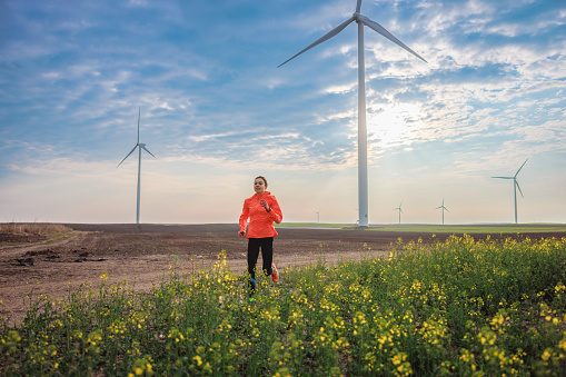 Young woman running in the field by the crops in rural area with wind turbines under the cloudy sky, active and healthy lifestyle