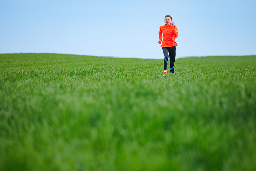 Young woman in sports outfit running through the grass in the meadow under the clear sky, active and healthy lifestyle