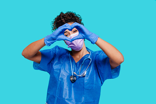 Nurse wears stethoscope and blue medical protective sterile gloves, does heart shape symbol with hands, isolated over blue background. Health care, medicine, protection, hospital concept.
