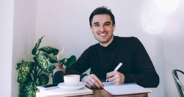 istock Happy young man writing essay while sitting in cafeteria with cup of coffee 1444171211