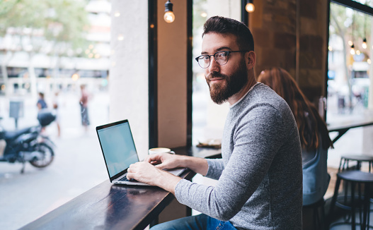 Side view of smiling bearded male remote employee in casual clothes and eyeglasses typing on netbook while sitting in cafeteria in afternoon and looking at camera