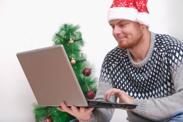 Man with laptop working, studying remotely new year 2023, Christmas. Man in Santa hat on background Christmas tree, garlands, gifts. Remote work. Surfing Internet. Man congratulates on holiday online. hombre con computadora portátil trabajando, estudiando de forma remota año nuevo 2023, navidad. hombre con sombrero de santa en el árbol de navidad de fondo, guirnaldas, regalos. trabajo remoto. navegación por internet. hombres felicita en vacaciones e