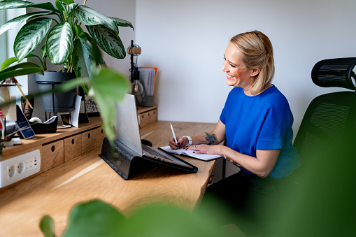 Young adult blond woman in blue blouse sitting at the desk in home office and using a laptop and a note pad.