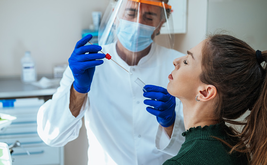 Mature male doctor wearing protective equipment and using cotton swab to test his female patient on possible coronavirus infection at medical clinic.