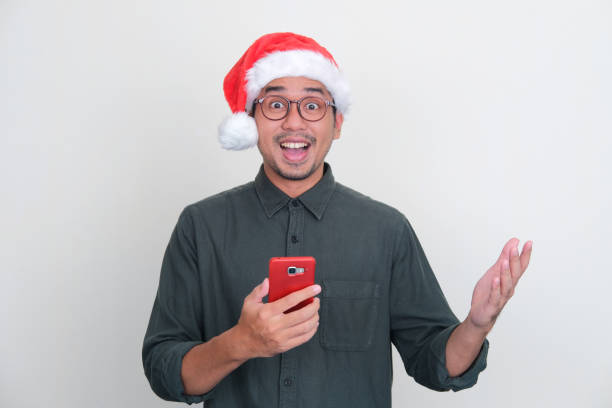 istock Asian man wearing Christmas hat looking camera with surprised expression while holding handphone 1444181384